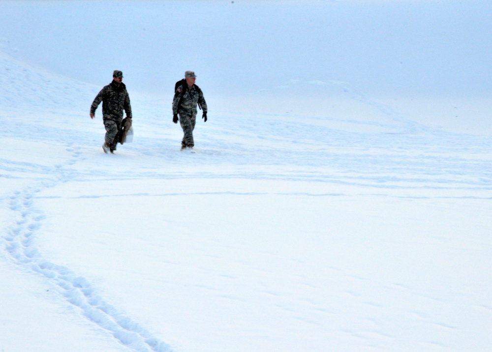 Two military personnel in camouflage uniforms are walking across a snow-covered open terrain. Their footprints are visible trailing behind them in the snow. The environment appears cold and overcast with low visibility conditions.