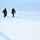 Two military personnel in camouflage uniforms are walking across a snow-covered open terrain. Their footprints are visible trailing behind them in the snow. The environment appears cold and overcast with low visibility conditions.