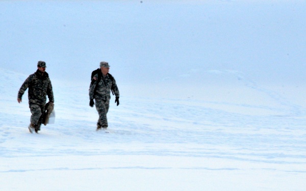 Two military personnel in camouflage uniforms are walking across a snow-covered open terrain. Their footprints are visible trailing behind them in the snow. The environment appears cold and overcast with low visibility conditions.