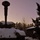 A military tank, heavily covered in snow, is photographed at dusk or dawn with a purple-pink sky in the background. The tank's gun barrel points upward prominently against the twilight sky. The setting appears to be an outdoor display or memorial site surrounded by snow-laden evergreen and deciduous trees.