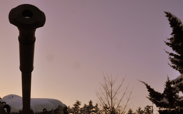 A military tank, heavily covered in snow, is photographed at dusk or dawn with a purple-pink sky in the background. The tank's gun barrel points upward prominently against the twilight sky. The setting appears to be an outdoor display or memorial site surrounded by snow-laden evergreen and deciduous trees.