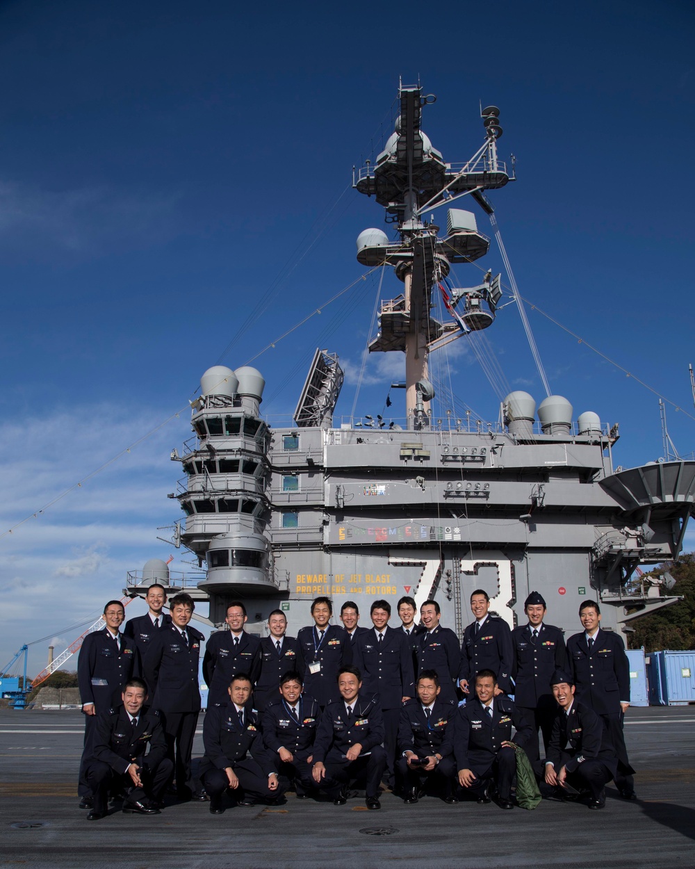 A group of approximately 20-25 military personnel in dark navy dress uniforms are posing for a group photograph on the flight deck of a large aircraft carrier. The island superstructure behind them displays the number '73' and the warning 'BEWARE OF JET BLAST, PROPELLERS, AND ROTORS,' identifying this as USS George Washington (CVN-73). The personnel appear to be members of the Japan Air Self-Defense Force (JASDF) based on their uniform style and insignia.