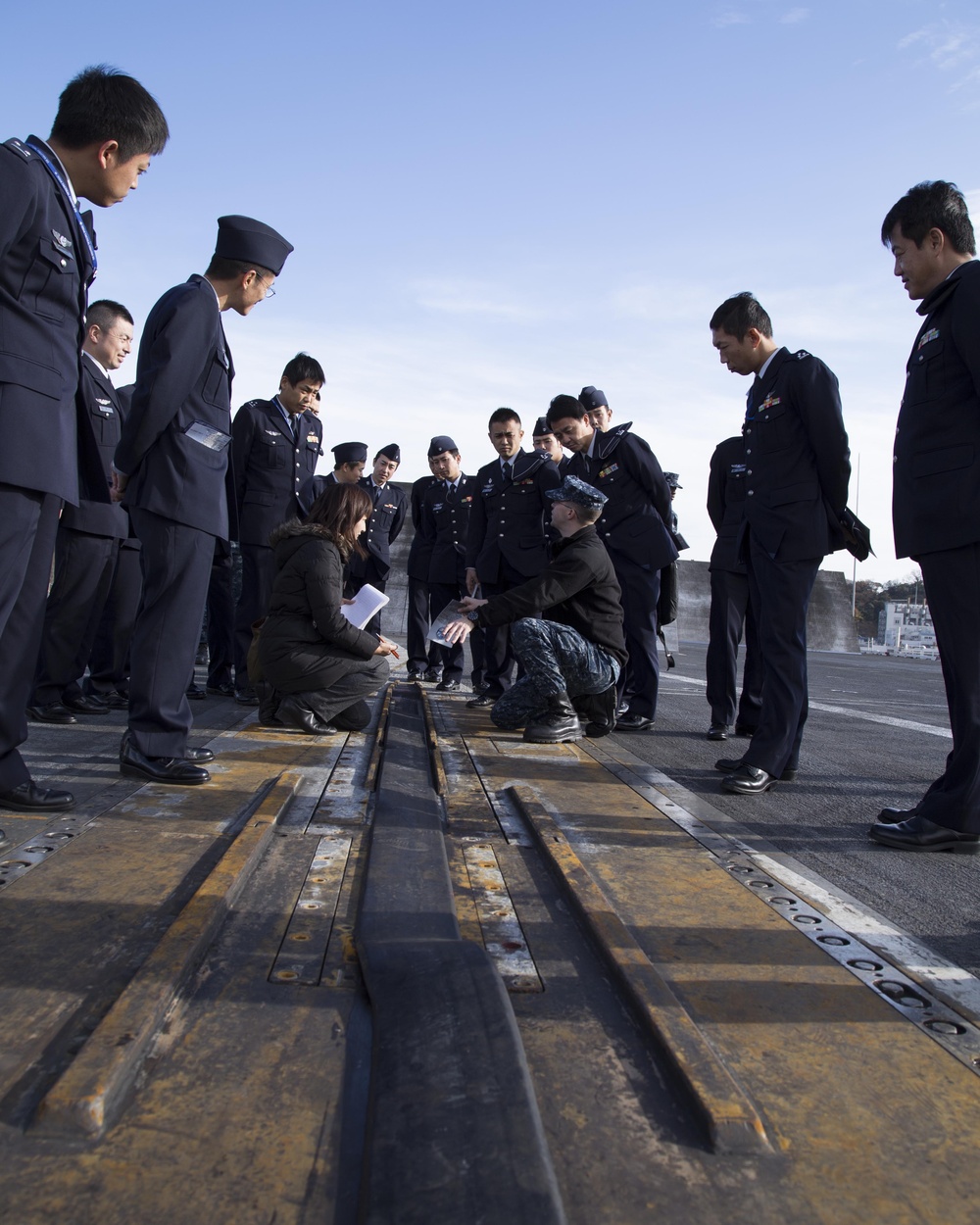 A group of uniformed military personnel, appearing to be Japanese Air Self-Defense Force officers, are gathered on what appears to be an aircraft carrier flight deck. A US Navy sailor in digital camouflage uniform is crouching down alongside a civilian interpreter to explain or demonstrate features of the catapult track system on the deck. This appears to be an official military exchange visit or tour.