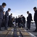 A group of uniformed military personnel, appearing to be Japanese Air Self-Defense Force officers, are gathered on what appears to be an aircraft carrier flight deck. A US Navy sailor in digital camouflage uniform is crouching down alongside a civilian interpreter to explain or demonstrate features of the catapult track system on the deck. This appears to be an official military exchange visit or tour.