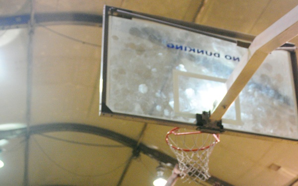 An indoor basketball game is in progress under a dome-shaped facility with a yellow court. A player in a red vest over a gray shirt is driving to the basket and attempting a layup while being contested by a defender in yellow. The backboard prominently displays a 'NO DUNKING' sign, visible in mirror-reversed text from this camera angle.