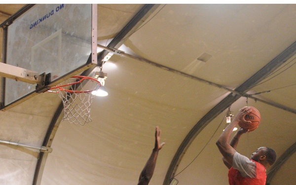 An indoor basketball game is being played inside what appears to be a military tent or dome facility, evidenced by the camouflage netting and temporary structure visible in the background. A player in a red jersey is driving to the basket for a layup attempt while a defender in a tan/khaki jersey contests the shot with an outstretched arm. Spectators are seated along the sidelines watching the game.
