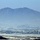 A wide-angle winter landscape showing a snow-covered plain with a small settlement or town cluster visible in the middle distance. A large, rugged mountain range dominates the background under a hazy blue sky. The scene appears to be viewed from an elevated vantage point across a flat agricultural or rural valley.