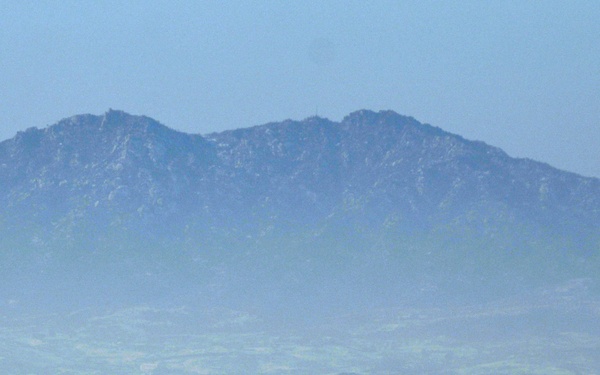 A wide-angle winter landscape showing a snow-covered plain with a small settlement or town cluster visible in the middle distance. A large, rugged mountain range dominates the background under a hazy blue sky. The scene appears to be viewed from an elevated vantage point across a flat agricultural or rural valley.