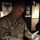 A U.S. Army soldier wearing ACU (Army Combat Uniform) is carefully handling what appears to be a Medal of Honor using white cotton gloves, likely in a museum or exhibit setting. The background displays exhibit panels referencing the Medal of Honor, SFC William S. Sitman, and a 'First Victory' display related to military history. A second pair of white-gloved hands is visible at the lower left, suggesting another person is also present.