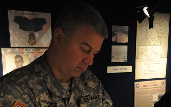 A U.S. Army soldier wearing ACU (Army Combat Uniform) is carefully handling what appears to be a Medal of Honor using white cotton gloves, likely in a museum or exhibit setting. The background displays exhibit panels referencing the Medal of Honor, SFC William S. Sitman, and a 'First Victory' display related to military history. A second pair of white-gloved hands is visible at the lower left, suggesting another person is also present.