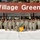A large group of U.S. Army soldiers in ACU (Army Combat Uniform) digital camouflage are posed for a formal unit photograph in front of a 'Village Green' facility sign. The group includes both male and female soldiers, with one soldier holding a unit guidon flag in green and yellow. The setting appears to be a military installation during winter, with snow visible on the ground.