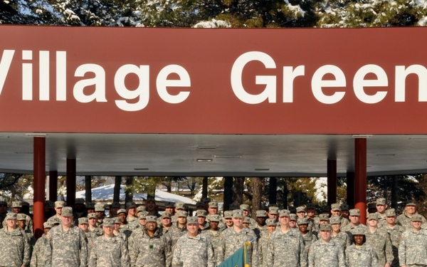 A large group of U.S. Army soldiers in ACU (Army Combat Uniform) digital camouflage are posed for a formal unit photograph in front of a 'Village Green' facility sign. The group includes both male and female soldiers, with one soldier holding a unit guidon flag in green and yellow. The setting appears to be a military installation during winter, with snow visible on the ground.