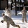 Three military personnel in US Army ACU (Army Combat Uniform) camouflage uniforms are engaged in a snowball fight in a snow-covered outdoor area. One soldier in the foreground is mid-throw, having just released a snowball that is visibly exploding in a spray of snow. A third soldier stands in the background with arms crossed, observing the activity.
