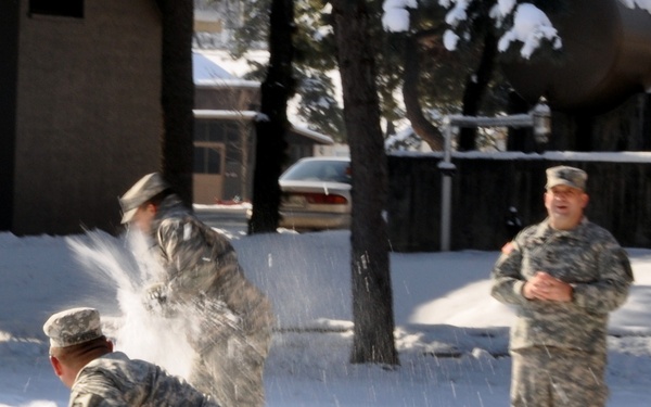 Three military personnel in US Army ACU (Army Combat Uniform) camouflage uniforms are engaged in a snowball fight in a snow-covered outdoor area. One soldier in the foreground is mid-throw, having just released a snowball that is visibly exploding in a spray of snow. A third soldier stands in the background with arms crossed, observing the activity.