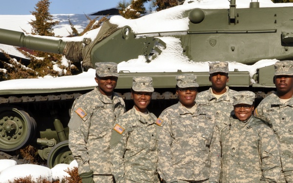 Five U.S. Army soldiers in ACU (Army Combat Uniform) digital camouflage are posing together in front of a static display tank in a snowy outdoor setting. The group appears to be at a military installation or museum, with snow covering the ground, benches, and the tank behind them. The soldiers are smiling and appear to be in a casual group photo.