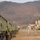 Three soldiers in US Army ACU (Army Combat Uniform) camouflage are walking across an open field at what appears to be a forward operating base or field exercise area. A row of olive-drab military trucks is lined up on the left side, while military tents, radar or communications dish equipment, and barbed wire perimeter fencing are visible in the background. The setting appears to be in a mountainous region during late autumn or winter, based on the bare deciduous trees on the hillside.