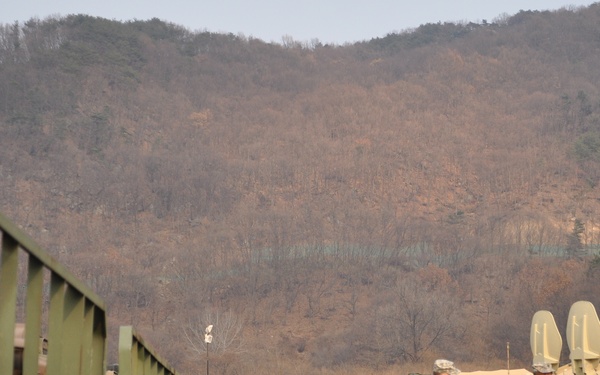 Three soldiers in US Army ACU (Army Combat Uniform) camouflage are walking across an open field at what appears to be a forward operating base or field exercise area. A row of olive-drab military trucks is lined up on the left side, while military tents, radar or communications dish equipment, and barbed wire perimeter fencing are visible in the background. The setting appears to be in a mountainous region during late autumn or winter, based on the bare deciduous trees on the hillside.