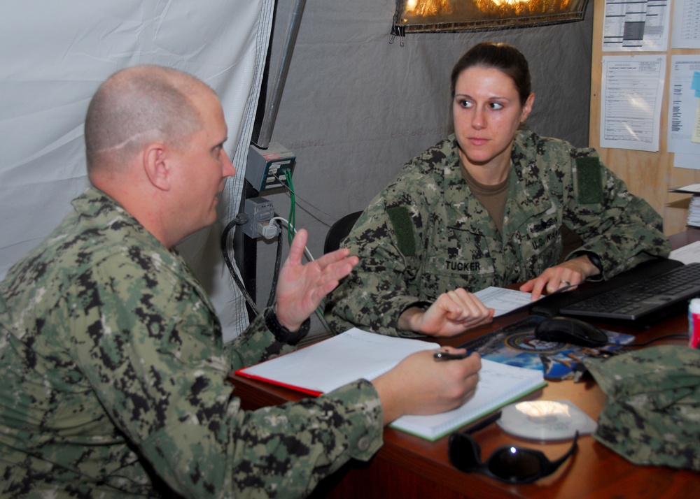 Two U.S. Navy personnel in NWU (Navy Working Uniform) digital camouflage are engaged in a meeting or briefing at a desk inside what appears to be a military field tent or temporary facility. The female sailor, identified by her name tape as 'TUCKER,' is seated across from a male sailor who appears to be speaking and gesturing. Documents, a laptop, and other office materials are visible on the desk.