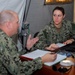 Two U.S. Navy personnel in NWU (Navy Working Uniform) digital camouflage are engaged in a meeting or briefing at a desk inside what appears to be a military field tent or temporary facility. The female sailor, identified by her name tape as 'TUCKER,' is seated across from a male sailor who appears to be speaking and gesturing. Documents, a laptop, and other office materials are visible on the desk.