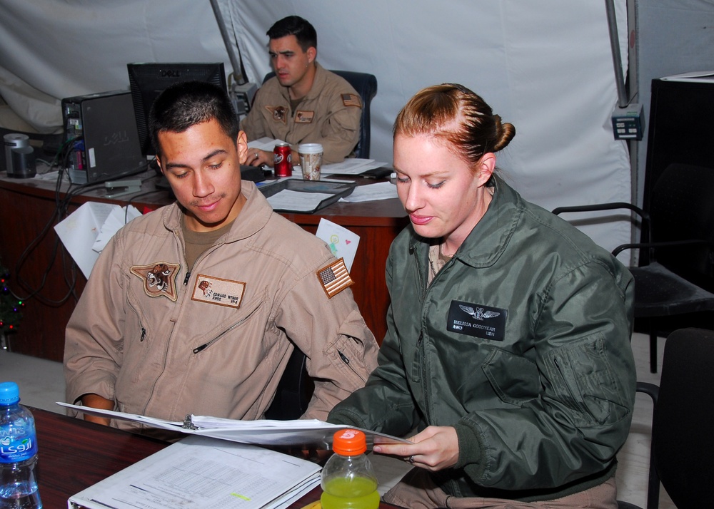 Three military personnel are working in what appears to be a forward operating base or field operations tent. Two individuals in the foreground — one in a tan flight suit and one in a green flight suit — are reviewing documents together at a desk. A third service member is visible in the background working at a computer workstation.