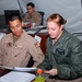 Three military personnel are working in what appears to be a forward operating base or field operations tent. Two individuals in the foreground — one in a tan flight suit and one in a green flight suit — are reviewing documents together at a desk. A third service member is visible in the background working at a computer workstation.