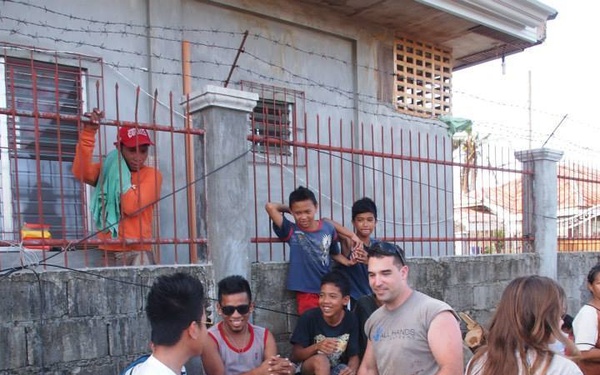 A group of volunteers and local community members, including many children, are gathered outside a concrete block building with orange metal security bars and barbed wire fencing. At least two individuals are wearing shirts identifying them as volunteers, suggesting this is a humanitarian or disaster relief effort. The scene appears to take place in a Southeast Asian community, likely the Philippines.