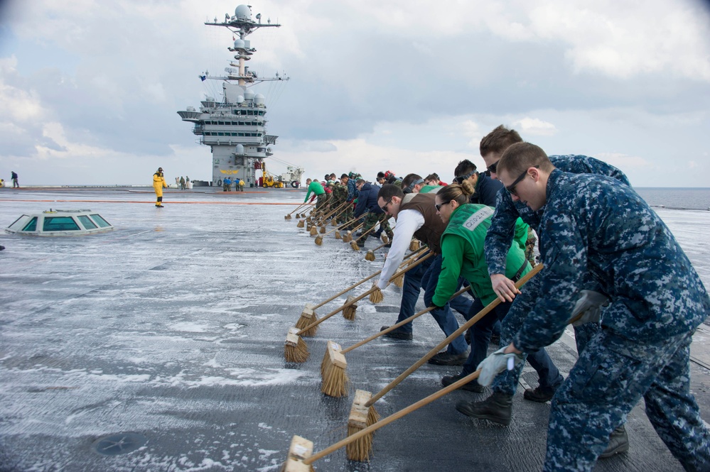 A large group of personnel, including U.S. Navy sailors in blue digital camouflage uniforms and civilians in casual clothing, are scrubbing the flight deck of an aircraft carrier using long-handled brooms with soapy water. The carrier's island superstructure with radar and communication equipment is visible in the background. This appears to be an organized deck-cleaning evolution, possibly during a distinguished visitor or Tiger Cruise event given the mix of military and civilian participants.