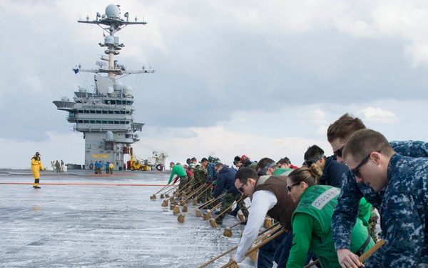 A large group of personnel, including U.S. Navy sailors in blue digital camouflage uniforms and civilians in casual clothing, are scrubbing the flight deck of an aircraft carrier using long-handled brooms with soapy water. The carrier's island superstructure with radar and communication equipment is visible in the background. This appears to be an organized deck-cleaning evolution, possibly during a distinguished visitor or Tiger Cruise event given the mix of military and civilian participants.