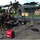 A group of military personnel in camouflage uniforms are gathered around a laptop computer in an outdoor staging area, operating what appears to be a portable satellite communications system. The setup includes a deployable satellite dish, ruggedized equipment cases, and a Honda EU2000i portable generator providing power. The scene suggests a forward operating base or disaster relief communications setup, with military tents, supply crates, and palletized cargo visible in the background.
