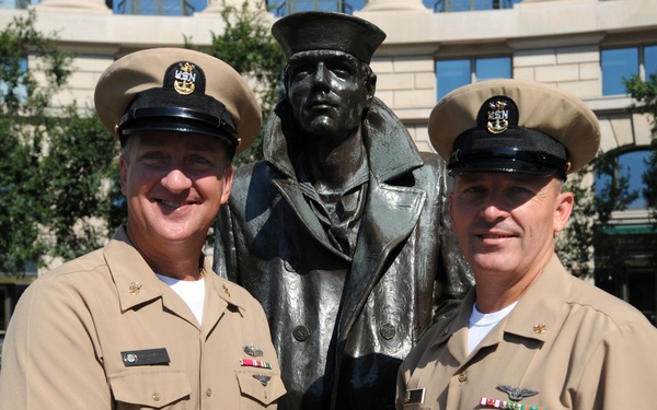 Two U.S. Navy senior enlisted personnel in khaki service uniforms are shaking hands in front of a bronze military statue. The setting appears to be an outdoor plaza, likely the U.S. Navy Memorial in Washington, D.C., based on the curved building and plaza design visible in the background. Both individuals wear covers (caps) with USN insignia and display ribbons and warfare qualification badges on their uniforms.