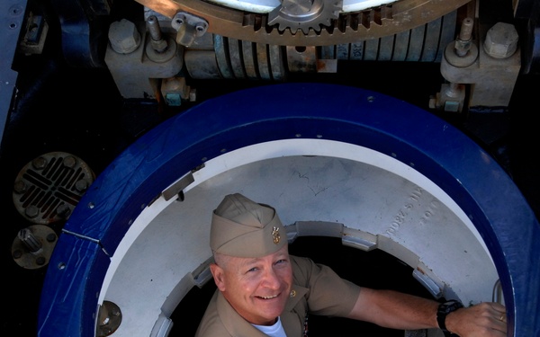 A smiling military officer in tan/khaki service uniform and garrison cap is emerging through a circular hatch, photographed from above. The hatch appears to be on a naval vessel or military platform, featuring a blue-rimmed circular opening with mechanical components visible around it. The individual appears to be climbing up through the access hatch and looking upward toward the camera.