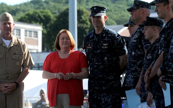 A military ceremony is taking place outdoors at what appears to be a U.S. Navy installation. A senior officer in tan/khaki dress uniform stands alongside a civilian woman in a red top, facing a formation of Navy personnel in blue digital camouflage uniforms. Several of the sailors in formation are holding certificates or documents, suggesting an awards or graduation ceremony.