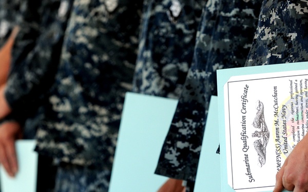 A row of United States Navy personnel in Navy Working Uniform (NWU) digital camouflage are standing at attention holding certificates. The closest certificate is visibly identified as a 'Submarine Qualification Certificate' issued to 'QMFN(SS) Aaron M. McCutcheon' of the United States Navy. This appears to be a military qualification or graduation ceremony recognizing submarine service achievements.