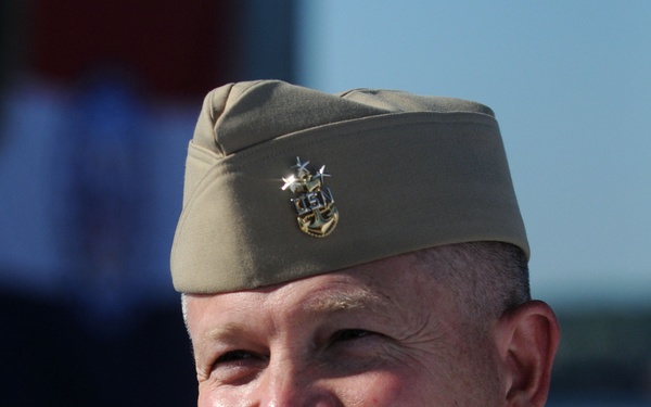 A middle-aged male in U.S. Navy enlisted uniform is smiling broadly, appearing to be at an outdoor military event or ceremony. He is wearing the khaki service uniform with collar insignia and a garrison/service cap bearing an enlisted rating badge. The background is blurred but suggests an outdoor setting with blue sky visible.