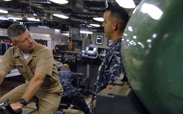 Two U.S. Navy personnel are visible in what appears to be a shipboard fitness area or hangar bay converted for exercise use. One sailor in khaki service uniform is seated on a stationary exercise bike, while another in Navy Working Uniform (NWU) digital camouflage stands nearby in conversation. The space features exercise equipment including treadmills and bikes, with industrial ship infrastructure visible in the background.