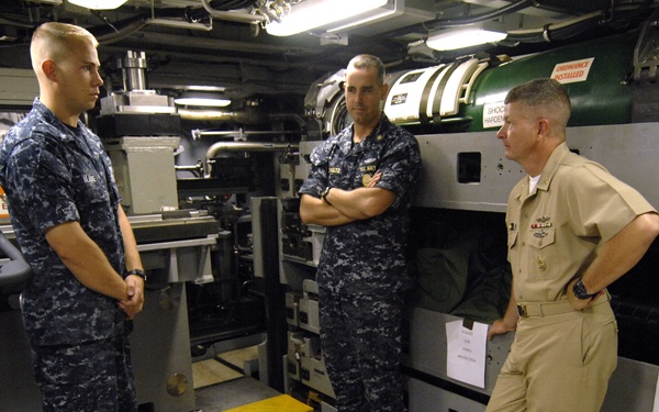 Three U.S. Navy personnel are gathered in what appears to be a submarine torpedo room that has been partially converted into a fitness area. Two sailors are wearing Navy Working Uniform (NWU) digital camouflage, while a third is in khaki service uniform, suggesting a senior officer conducting an inspection or walkthrough. The space contains exercise equipment alongside torpedo tubes and military hardware.