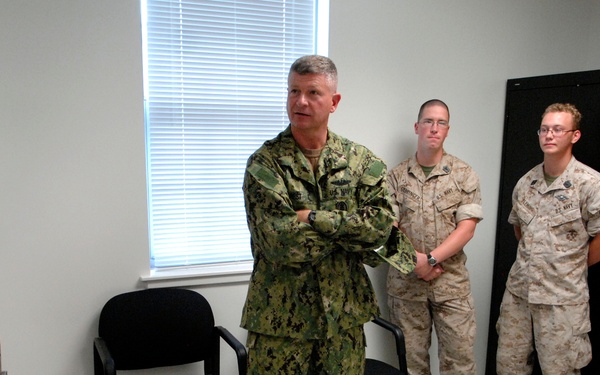 A group of military personnel in a sparse indoor room, appearing to be in a briefing or meeting setting. The central figure, wearing U.S. Navy NWU (Naval Working Uniform) camouflage, stands with arms crossed in a position of authority. Surrounding personnel wear a mix of U.S. Navy and U.S. Marine Corps MARPAT desert/woodland camouflage uniforms.