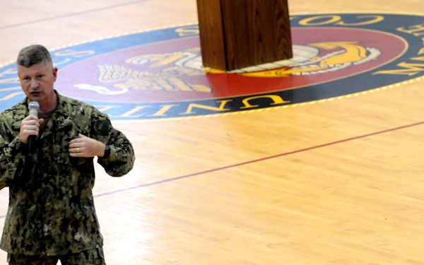 A military officer in digital camouflage uniform is speaking into a handheld microphone on the floor of a gymnasium. The gym floor features a United States Marine Corps emblem/seal in the center court area. The setting appears to be an official military address or all-hands meeting.