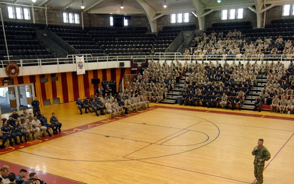 A large military assembly is taking place inside a Marine Corps gymnasium/arena, with hundreds of service members in desert camouflage and Navy uniforms seated around the basketball court. A single speaker in military uniform stands on the court floor addressing the assembled troops, with a podium visible to the right. The United States Marine Corps emblem is visible on the court floor and on banners/seals around the facility.