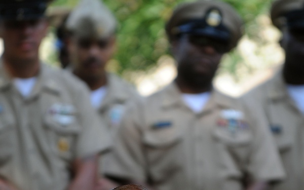 An elderly man seated in a wheelchair is positioned in the foreground, wearing a straw fedora hat, glasses, a blue dress shirt, and a tan sweater vest. Behind him stand several individuals in tan/khaki military-style uniforms with rank insignia and name tags, appearing to be at an outdoor ceremony or formal event. The composition suggests the elderly man may be a veteran being honored at a military or commemorative gathering.