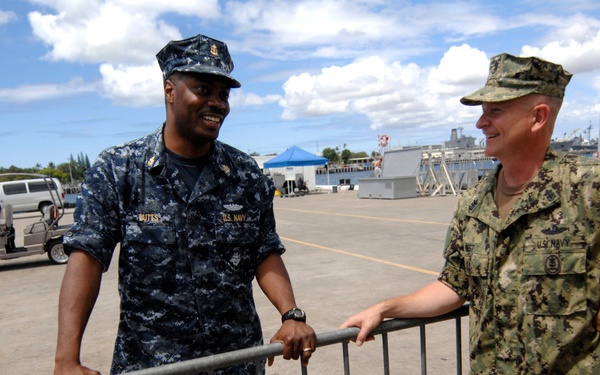Two U.S. Navy personnel are conversing at what appears to be a naval base pier or dock area. One sailor is wearing the Navy Working Uniform (NWU) Type I blue digital camouflage, while the other is wearing NWU Type III green/woodland camouflage. Naval vessels are visible in the background harbor.