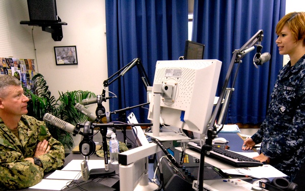 Two U.S. Navy personnel in digital camouflage uniforms are in what appears to be a military radio broadcast studio. One individual is seated at a desk with microphones and broadcasting equipment, while the other stands across the desk. An 'Air Force' framed poster with an eagle is visible on the wall in the background.