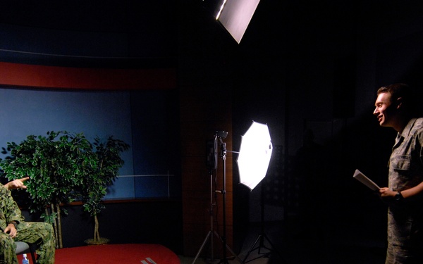 A U.S. Navy officer in camouflage uniform is seated on a raised red-carpeted studio platform, appearing to be interviewed or filmed in a professional broadcast studio setting. A civilian or crew member stands to the right holding papers, likely conducting the interview or directing the session. The studio is equipped with professional lighting equipment including an octabox softbox on a stand.