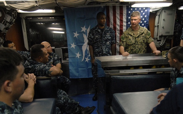 A senior military officer in tan/khaki uniform addresses a group of Navy sailors in a shipboard compartment, likely a mess deck or briefing room. The sailors are wearing Navy Working Uniform (NWU) blue digital camouflage pattern. Two flags are visible in the background: the American flag and a blue flag with white stars and a figure, likely a command or fleet flag.