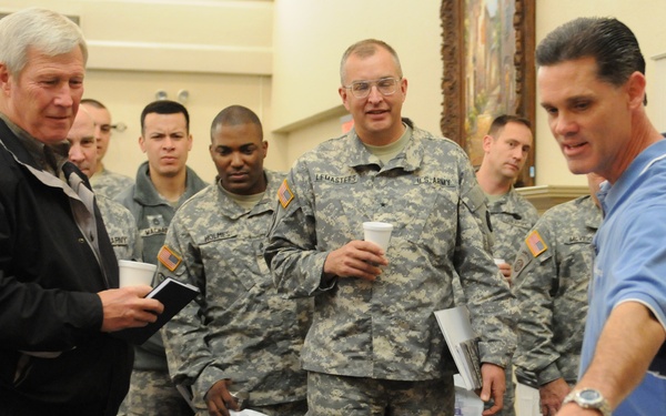 A group of U.S. Army soldiers in ACU (Army Combat Uniform) camouflage and two civilians are gathered around a table with equipment during what appears to be a briefing or demonstration. A man in a blue polo shirt is presenting or explaining equipment to the group. The setting appears to be an indoor conference or meeting room.