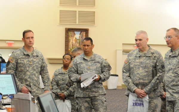 A group of U.S. Army soldiers in ACU (Army Combat Uniform) camouflage are gathered at what appears to be a military job fair, transition assistance event, or administrative processing station. A civilian presenter or representative stands behind a table equipped with multiple computer monitors and laptops. The setting appears to be a conference or banquet hall.