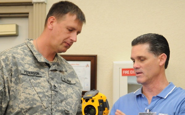 A U.S. Army soldier in ACU (Army Combat Uniform) is being shown a yellow thermal imaging camera by a civilian representative wearing a blue polo shirt and an exhibitor badge. The civilian, whose badge indicates affiliation with 'EN TEST,' appears to be demonstrating or explaining the device's features. The setting appears to be an indoor exhibition or trade show environment.