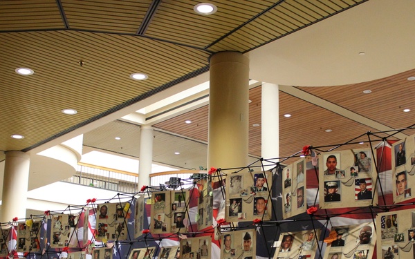 A large memorial display is set up inside what appears to be a convention center or public building, featuring numerous photographs of military personnel mounted on American flag-themed panels. The exhibit appears to be a tribute to fallen or serving military members, with red poppy or ribbon decorations adorning the display frames. A single person can be seen on the left side viewing the memorial.
