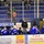 A hockey team bench labeled 'VISITORS' with players wearing blue 'ICEMEN' jerseys during what appears to be a game or practice session at an indoor ice rink. A coach in a black hoodie and cap stands behind the bench observing the action. Sparse spectators are visible in the blue stadium seating behind the glass.