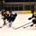 An ice hockey game is in progress featuring players from two teams: one in black and gold 'ARMY' jerseys and one in blue jerseys. A player in blue is controlling the puck while being challenged by opposing players wearing black and gold uniforms. The action takes place in an indoor ice rink with rinkside advertising banners visible in the background, including a 'Special Olympics' banner.